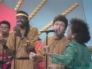 Grupo de vocalistas en un programa de televisión de estética retro, varios cantantes frente a micrófonos con vestuario colorido —una mujer con blusa verde brillante y hombres con prendas en tonos tierra y una cinta en la cabeza— sobre un fondo con luces decorativas y paneles geométricos, Qué ventajas y desventajas tiene la yuca: beneficios y riesgos