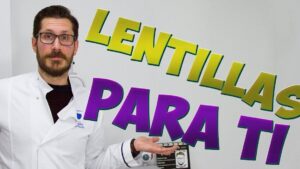 Optometrista con bata blanca y gafas sostiene la mano abierta mientras posa junto a modelos y material ocular delante de un fondo con grandes letras coloridas que anuncian lentillas, Ventajas y desventajas de las lentillas: todo lo que debes saber.