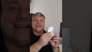 Hombre sonriente en primer plano sentado en un escritorio con un monitor al fondo sostiene frente a la cámara un estuche pequeño y brillante, escena clara y accesible que ilustra 'Ventajas y desventajas de los ordenadores: lo que debes saber'.