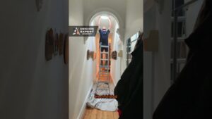 Persona de pie sobre una escalera en un pasillo estrecho de paredes blancas y suelo de madera, ajustando una claraboya redonda mientras una lona protege el suelo y cuadros decoran las paredes, Ventajas y desventajas del último piso: descubre si es para ti
