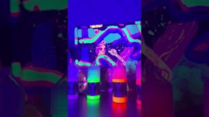 Varias botellas pequeñas con líquidos fluorescentes en tonos verde, naranja y rosa que brillan bajo luz negra frente a un fondo abstracto y psicodélico, imagen que ilustra Luz ultravioleta en alimentos: ventajas y desventajas esenciales y ofrece una descripción accesible centrada en envases, colores neón y reflejos para mejorar SEO y comprensión para lectores con discapacidad visual.