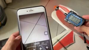 Manos sosteniendo un teléfono con la cámara abierta que muestra una lectura digital y, junto a ello, otra mano que sujeta un tacómetro portátil sobre la hélice de un avión RC de espuma rojo y blanco sobre suelo de baldosas, ilustrando Ventajas y desventajas de un medidor de rpm clave.