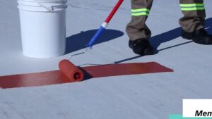 Trabajador con pantalones y botas de seguridad aplicando con rodillo un recubrimiento rojo sobre una cubierta plana blanca junto a un cubo, imagen que sirve como ilustración de Membranas de poliuretano ventajas y desventajas clave.