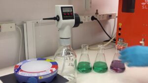 Imagen de una bancada de laboratorio con un dispensador automático, tres frascos Erlenmeyer con soluciones verdes y violeta, un agitador magnético y una mano con guante azul manipulando materiales de titulación, útil como ilustración para Ventajas y desventajas del método Kjeldahl en análisis de nitrógeno y optimizada para accesibilidad web y SEO.