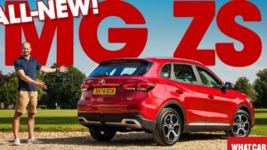 Un hombre presenta un MG ZS rojo visto de tres cuartos por detrás, aparcado sobre grava frente a un césped y árboles con grandes letras rojas promocionales en el fondo, mostrando detalles de las llantas, el paragolpes trasero y las líneas de la carrocería, Ventajas y desventajas de MG ZS, el SUV híbrido ideal para ti.