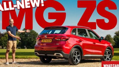 Un hombre presenta un MG ZS rojo visto de tres cuartos por detrás, aparcado sobre grava frente a un césped y árboles con grandes letras rojas promocionales en el fondo, mostrando detalles de las llantas, el paragolpes trasero y las líneas de la carrocería, Ventajas y desventajas de MG ZS, el SUV híbrido ideal para ti.