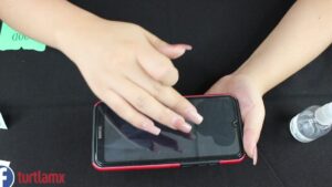Manos femeninas con uñas esmaltadas presionan y alinean un protector sobre la pantalla de un teléfono móvil sobre una superficie negra junto a un frasco con atomizador, Mica de cerámica ventajas y desventajas: ¿es la mejor opción?