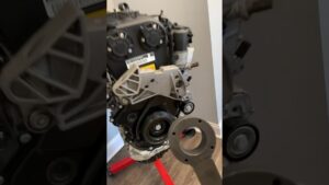 Vista frontal y detallada de un motor TSI desmontado colocado sobre un soporte rojo en un taller doméstico, con la carcasa delantera, poleas, correa y soporte de motor visibles y una herramienta con abertura circular en primer plano para inspección y montaje, Ventajas y desventajas del motor tsi: ¿vale la pena?