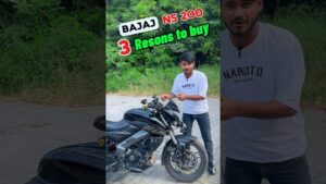 Hombre joven con camiseta blanca con el texto "Naruto Shippuden" presenta y señala una motocicleta deportiva negra Bajaj NS 200 aparcada sobre pavimento frente a vegetación, con texto superpuesto promocional que resalta motivos para comprar; Ventajas y desventajas de la NS 200: ¿es la moto ideal?