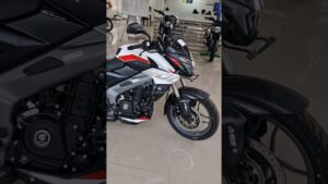 Motocicleta naked deportiva blanca con detalles rojos y negros expuesta en un concesionario, vista frontal lateral derecha que muestra el faro agresivo, el manillar con retrovisor, el depósito, el motor a la vista, la llanta delantera con disco y la pegatina ABS sobre suelo pulido con otras motos y material informativo al fondo, Ventajas y desventajas de la Rouser NS 200: ¿vale la pena?