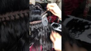 Vista posterior de una instalación de extensiones con manos de la estilista sujetando el cabello oscuro con pinzas mientras se aprecia una hilera de pequeños nudos en la raíz y mechas onduladas que se incorporan al cuero cabelludo — Extensiones de nudo brasileño ventajas y desventajas para ti