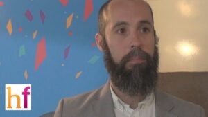 Hombre con barba oscura y traje claro en primer plano sentado frente a una pared azul decorada con formas geométricas de colores y un pequeño logotipo en la esquina inferior izquierda, imagen neutra y accesible apropiada para acompañar Ventajas y desventajas del ocio digital: impacto real y sorprendente