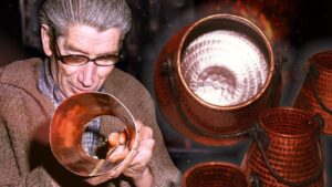 Hombre mayor con gafas inspeccionando una lámina de cobre mientras se aprecian varias ollas de cobre martilladas con interior pulido y asas en un taller artesanal, evocando técnicas tradicionales de manufactura y cuidado de utensilios; Ollas de cobre ventajas y desventajas: ¿valen la pena?