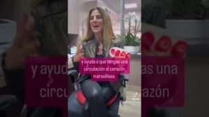 Fotograma vertical de un vídeo en formato entrevista que muestra a una mujer de cabello largo y chaqueta de cuero sentada y gesticulando en una sala con plantas y una orquídea, con un recuadro rosa con texto que dice "y ayuda a que tengas una circulación al corazón maravillosa", y Ventajas y desventajas del óxido nitroso: descubre sus efectos clave.