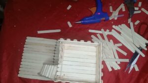 Vista cenital de un proyecto de manualidades sobre tela roja con palitos de helado organizados en una base rectangular y una caja en construcción, pistola de pegamento caliente con luz roja, tijeras y palitos sueltos dispersos que describen materiales, herramientas y pasos DIY para accesibilidad y SEO; Ventajas y desventajas de las pajas: efectos reales y sorprendentes