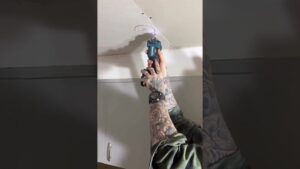 Manos tatuadas sosteniendo una herramienta eléctrica mientras realizan un corte circular en una placa de yeso del techo junto a tornillos visibles sobre un mueble, ilustrando Ventajas y desventajas del panel de yeso para tu proyecto y destacando aspectos de instalación, corte y acabado para accesibilidad y optimización.