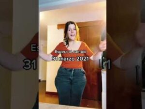 Mujer joven sonriente de pie en un interior con luz cálida, brazos abiertos delante de una puerta, viste camiseta naranja y pantalones vaqueros en un encuadre vertical con texto superpuesto, Ventajas y desventajas de no cenar y su impacto en tu salud
