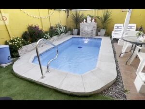 Piscina de fibra de forma rectangular con agua azul y borde de hormigón, barandilla metálica de acceso, fuente decorativa contra una pared amarilla rodeada de plantas y muebles de jardín, Ventajas y desventajas piscinas de fibra: ¿es la opción ideal?