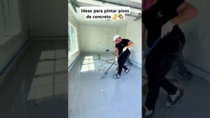 Obrero con mascarilla, guantes y zapatillas aplica con rodillo una capa de resina epóxica gris sobre el suelo de una habitación o garaje iluminado por amplias ventanas mientras los zócalos están protegidos con cinta, mostrando el proceso de nivelado y el acabado brillante que ilustra Ventajas y desventajas de piso epóxico: ¿es la mejor opción para ti?.