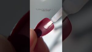 Primer plano de una mano que sostiene una uña postiza color vino tinto mientras un pincel aplica adhesivo transparente en su base, imagen que ilustra Uñas press on ventajas y desventajas: ¿son la mejor opción para ti?