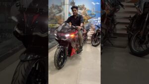 Hombre sentado sobre una motocicleta deportiva negra y roja en el interior de un concesionario con varias motos al fondo, con las luces delanteras encendidas y postura de prueba que sugiere una reseña visual, Pulsar rs 200 ventajas y desventajas: ¿vale la pena?