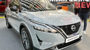 Fotografía frontal en tres cuartos de un Nissan Qashqai plateado expuesto en un salón, con parrilla V‑Motion y emblema Nissan bien visibles, faros LED delgados, llantas de aleación, reflejos del techo y personas difuminadas al fondo — Ventajas y desventajas Nissan Qashqai 2024 que debes conocer sobre diseño y acabados.