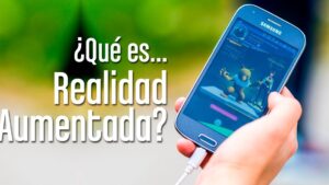 Primer plano de una mano que sostiene un smartphone Samsung conectado a un cable de carga y muestra un juego de realidad aumentada con un Pokémon y su entrenador en la pantalla, sobre un fondo exterior desenfocado y con un gran texto blanco a la izquierda que pregunta "¿Qué es... Realidad Aumentada?", imagen ideal para contenidos y accesibilidad que ilustra Ventajas y desventajas de AR y VR: descubre sus impactos y retos clave.