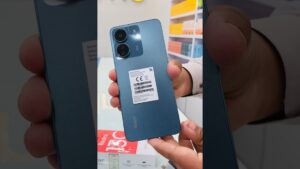 Mano sostiene un Xiaomi Redmi 13c azul verdoso mostrando la trasera con dos módulos de cámara circulares, acabado mate y etiqueta blanca visible sobre la carcasa, con la caja del producto y estantería de tienda desenfocadas al fondo, imagen ideal para reseñas y comparativas: Xiaomi Redmi 13c ventajas y desventajas para decidir su compra