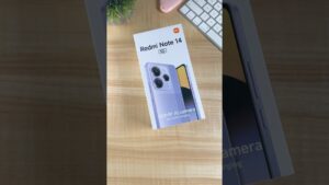 Caja blanca del Redmi Note 14 5G apoyada sobre un escritorio de madera junto a un teclado blanco, una funda rosa y una planta decorativa, con la ilustración del teléfono y sus grandes lentes visibles, pensada para imágenes de unboxing y reseñas; Ventajas y desventajas del Redmi 14 cc: ¿vale la pena?