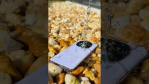Teléfono blanco con la palabra Redmi en la carcasa apoyado sobre guijarros mientras un chorro de agua salpica el módulo circular de la cámara, imagen clara y descriptiva ideal para artículos y comparativas como Xiaomi Redmi Note 13 ventajas y desventajas para decidir su compra.