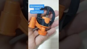 Primer plano de una mano sosteniendo un microaspersor plástico naranja y negro con base roscada y rotor giratorio visible para sistemas de riego, Ventajas y desventajas riego por aspersión: ¿es ideal para ti?