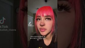Retrato vertical en primer plano de una persona joven con flequillo rosa y mechones oscuros, pestañas largas y maquillaje visible, piercings en la nariz, expresión pensativa frente a una puerta y un reloj difuminados y con marca de agua de TikTok visible, Sa de cv ventajas y desventajas para elegir la mejor opción