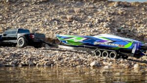 Una camioneta pickup negra remolca un bote de carreras azul, verde y morado sobre un remolque de triple eje en una orilla pedregosa junto al agua — Ventajas y desventajas de S de RL: ¿es la mejor opción para ti?