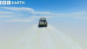 Un todoterreno solitario recorre una vasta llanura blanca que se funde con el horizonte bajo un cielo azul con nubes y el logotipo de BBC Earth en la esquina superior izquierda, evocando Ventajas y desventajas de la explotación del salar de Uyuni al sugerir contrastes entre turismo, extracción de recursos y la fragilidad del paisaje salino.