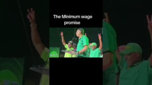 Orador en un podio con micrófono y varias personas vestidas de verde levantando el gesto de victoria en un acto público con un cartel en inglés sobre promesa de salario mínimo, Ventajas y desventajas de la UMA en tus finanzas y derechos