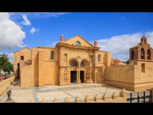 Fachada monumental de una catedral colonial en piedra amarilla con portada ornamentada de arcos y columnas, campanario lateral, amplio atrio pavimentado y detalles escultóricos bajo un cielo azul brillante, texto alternativo optimizado para accesibilidad y SEO que incluye Ventajas y desventajas de la isla de Santo Domingo: secretos clave para orientar a visitantes y lectores sobre patrimonio histórico.