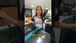 Una mujer en una tienda de electrodomésticos, con camiseta de marca, gesticula entre una lavadora y una secadora durante una demostración mientras en la pantalla aparece el texto la ropa, ilustrando de forma visual el tema Ventajas y desventajas secadora de ropa: ¿vale la pena usarla?