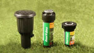 Tres cabezales emergentes de aspersores sobre césped, dos con etiqueta verde de la marca Rain Bird y uno negro de mayor tamaño fotografiados en primer plano para comparar diseño, materiales y conexiones — Sistema de irrigación colostomía: ventajas y desventajas clave