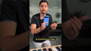 Hombre de pie en un interior sostiene un teléfono móvil azul frente a la cámara mientras varios móviles están alineados sobre una mesa y se aprecia decoración con reloj en la pared y subtítulos amarillos, descripción visual clara y optimizada para SEO y accesibilidad, Sistema operativo Linux ventajas y desventajas: descubre si es para ti