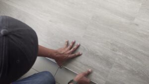 Persona con gorra y jeans arrodillada que ajusta con las manos una lama gris imitación madera de suelo SPC colocándola sobre baldosas, mostrando el proceso de instalación, el encaje y la textura para facilitar la evaluación, Ventajas y desventajas del sistema SPC: claves para decidir fácil