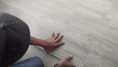 Persona con gorra y jeans arrodillada que ajusta con las manos una lama gris imitación madera de suelo SPC colocándola sobre baldosas, mostrando el proceso de instalación, el encaje y la textura para facilitar la evaluación, Ventajas y desventajas del sistema SPC: claves para decidir fácil