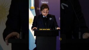 Persona de mediana edad con traje oscuro y gafas leyendo un discurso detrás de un atril con micrófonos y una bandera argentina de fondo, imagen apta para ilustrar análisis sobre Socialismo del siglo xxi ventajas y desventajas y sus impactos clave