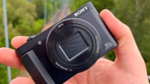 Mano sosteniendo una cámara compacta negra Sony con objetivo Zeiss y tapa de lente cerrada, agarre texturizado y marcado de zoom óptico treinta veces sobre un fondo exterior verde desenfocado, Sony DSC H200 ventajas y desventajas para decidir si es para ti.