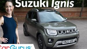 Mujer sonriente junto a un Suzuki Ignis verde oliva estacionado en un entorno urbano con valla de madera y árboles, imagen de reseña que invita a leer "Ventajas y desventajas del Suzuki Ignis: ¿vale la pena comprarlo?" resaltando diseño compacto, faros delanteros y carácter práctico del vehículo.