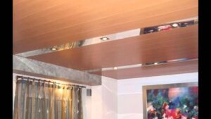 Sala interior con techo suspendido formado por paneles de apariencia madera y franjas metálicas reflectantes con focos empotrados, cortinas translúcidas y un cuadro abstracto en la pared, Techos de zinc ventajas y desventajas: ¿son la mejor opción?