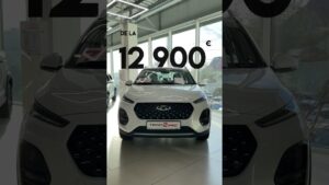 En un concesionario iluminado se muestra la vista frontal de un SUV compacto blanco con parrilla negra de panal, faros delgados, placa frontal que indica Tiggo 2 Pro y carteles de precio en la ventana; Tiggo 2 Pro ventajas y desventajas: ¿vale la pena este SUV?