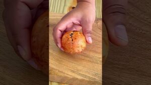 Primer plano de manos que sostienen una esfera redondeada de color dorado anaranjado con motas oscuras y una costura visible sobre una tabla de madera que resalta la textura jaspeada y el brillo superficial, Henna para el cabello ventajas y desventajas: ¿es ideal para ti?