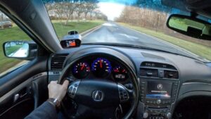 Vista interior desde la posición del conductor con la mano apoyada en el volante, panel de instrumentos iluminado con tacómetro y velocímetro, pantalla de navegación en la consola central, retrovisor y vista despejada de la carretera frontal, Acura TL 2005 ventajas y desventajas clave antes de comprar