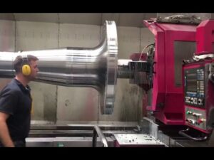 Operario con protección auditiva supervisa un gran torno CNC mientras maquila una pieza metálica cilíndrica brillante, con panel de control numérico a la derecha y refrigerante y virutas visibles en el área de mecanizado del taller industrial; Ventajas y desventajas del torno CNC: ¿vale la pena invertir?