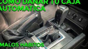 Primer plano del interior de un automóvil que muestra la palanca de cambios automática con funda de cuero y el freno de mano junto a la consola central con botones y superficies algo desgastadas, con texto verde sobre malos hábitos, Transmisión automática CVT ventajas y desventajas para ti